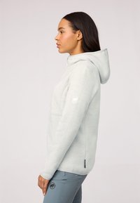 Felpa grigia chiara con zip e texture morbida, dotata di cappuccio e toppa con logo sul braccio, abbinata a pantaloni blu-grigi aderenti.