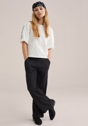 Jauna sieviete stāv, liecot rokas kabatās, valkājot baltu īsroku topu, melnas faktūras bikses, melnus zābakus un melnu rakstainu galvas apsēju.