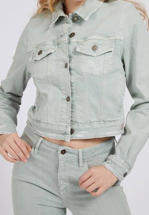 Frau trägt eine hellgrüne, kurze Jeansjacke mit Knopfleiste vorne und passende hoch taillierte Jeans, Hände in der Nähe von Taille und Hüfte.