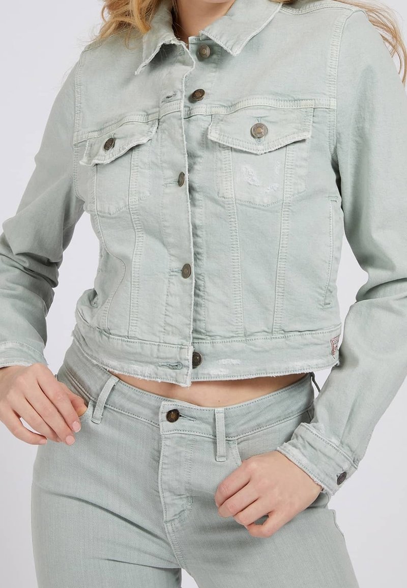 Femme portant une veste en denim courte vert clair avec fermetures boutonnées à l'avant et un jean taille haute assorti, mains près de la taille et des hanches.