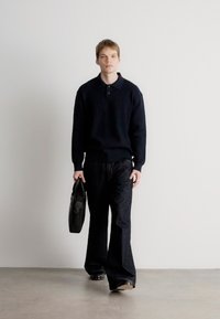 Pulôver preto de malha canelada, calças largas pretas e sapatos pretos. A carregar uma bolsa de couro preta. Fundo neutro.