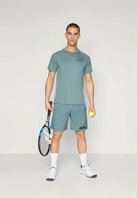 Junger Mann, der steht und in einer Hand einen Tennisschläger und in der anderen einen Tennisball hält. Er trägt passend türkisfarbene Sportkleidung und weiße Turnschuhe.