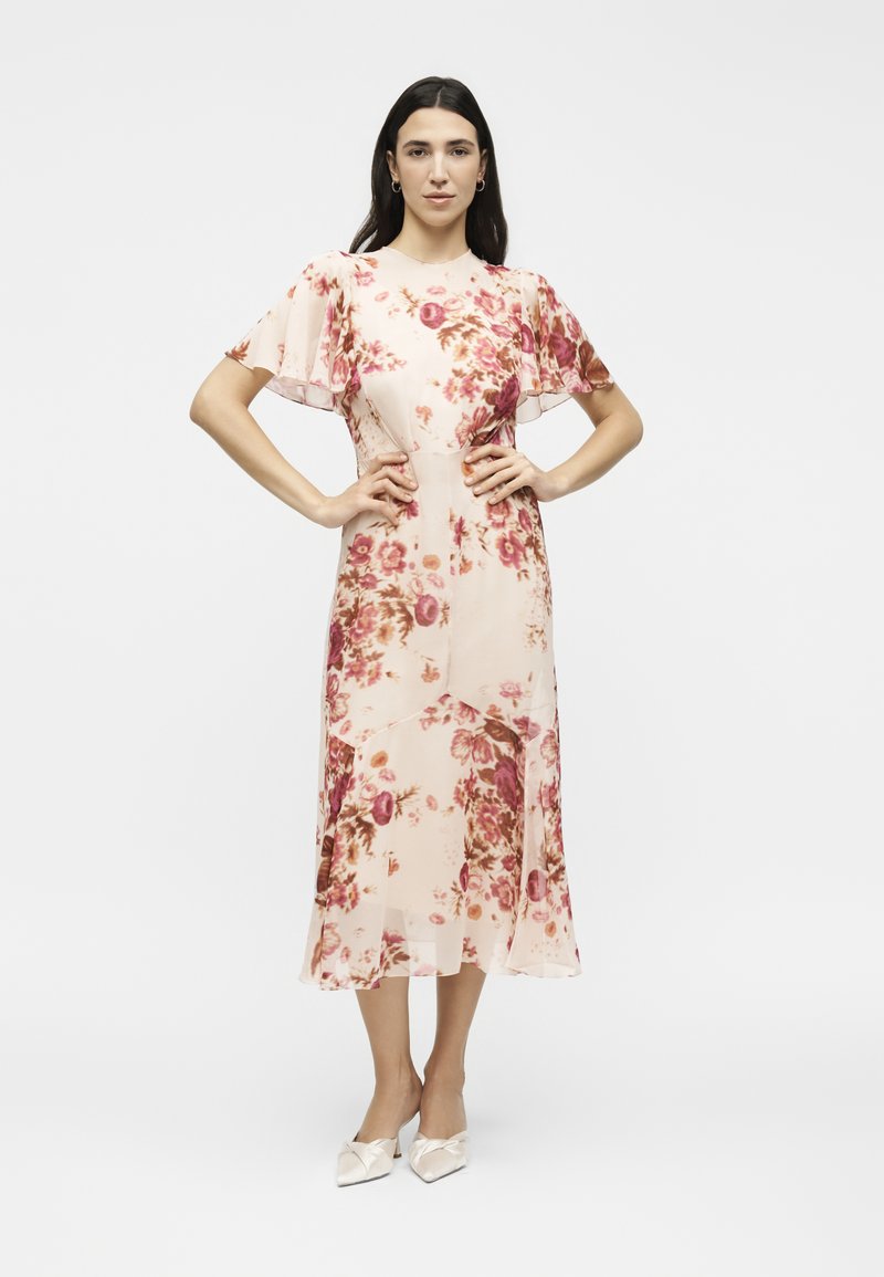 Frau steht mit den Händen in den Hüften, trägt ein hellrosa Midikleid mit rosa und braunen Blumenmustern und weiße spitze Schuhe.