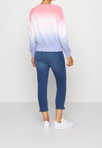 Persona che indossa un felpa ombre da rosa a viola, jeans blu cropped con tasche posteriori e sneakers bianche, in piedi contro uno sfondo semplice.