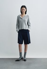 Hellgrauer gestrickter Pullover mit Kragen, kombiniert mit marineblauen plissierten Shorts und grauen gerippten Socken, dazu schwarze spitze High Heels.