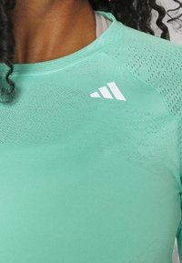 Mintgrünes Sportshirt aus glattem, dehnbarem Stoff. Hat einen runden Halsausschnitt und ein weißes Logo auf der linken Brust mit strukturierten Details.
