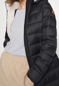 Sort dunjakke med hvid fleeceforing, quiltet design, lynlåslukning og et orange logo-patch på venstre ærme.