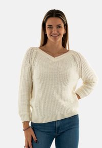 Pull en tricot crème à col en V, avec un motif texturé et des manches trois-quarts. Dispose de poignets et d'un ourlet côtelés. Associé à un jean bleu.