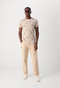 Mønstret kortærmet t-shirt i beige med farverige geometriske designs; parret med lyse beige bukser og hvide sneakers. Stilfulde solbriller.