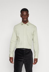 Chemise vert clair à manches longues avec col, fermeture à boutons à l'avant et tissu lisse ; associée à un pantalon noir texturé.