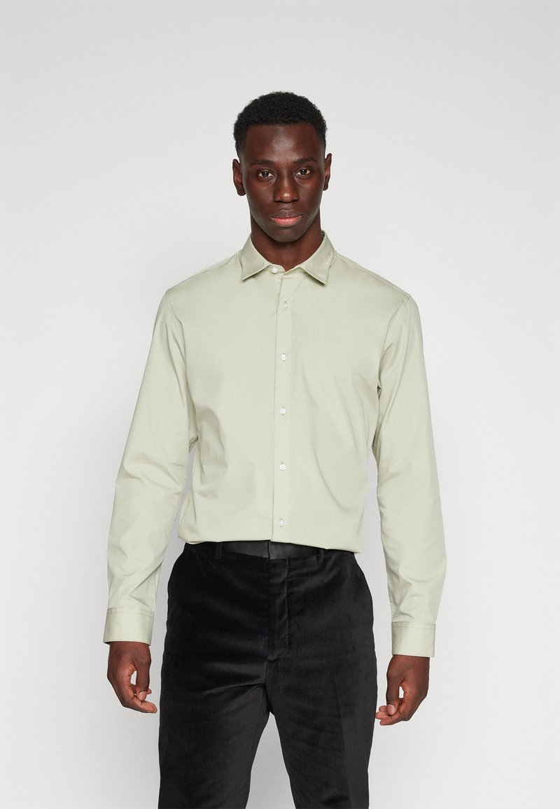 Chemise vert clair à manches longues avec col, fermeture à boutons à l'avant et tissu lisse ; associée à un pantalon noir texturé.