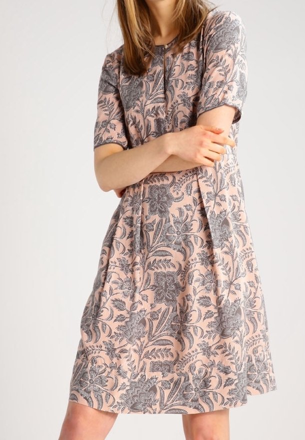Femme portant une robe rose mi-longue à manches courtes avec un motif floral gris, les bras croisés, debout devant un fond clair uni.