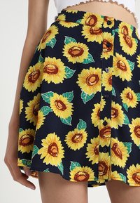 Navyblauwe button-up rok met een geel zonnebloem- en groen bladpatroon, gedragen met een witte crop top.