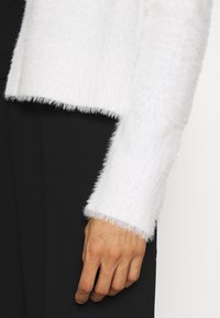 Main reposant sur le côté, portant un pull blanc duveteux à manches longues avec un pantalon noir sur un fond neutre.