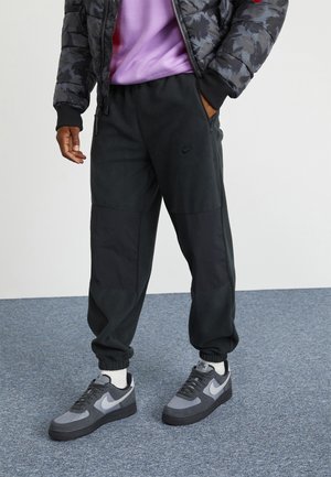 Mężczyzna noszący czarne spodnie dresowe Nike, szaro-czarne buty Nike, białe skarpetki, fioletową koszulkę oraz ciemną kamuflażową kurtkę puchową.