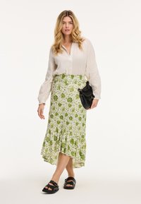 Witte, knoopsluiting blouse van textuurstof gecombineerd met een groene rok met bloemenpatroon, voorzien van een rucheszoom en zwarte sandalen.