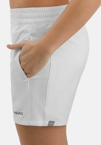 Mano descansando en el bolsillo lateral de pantalones cortos deportivos blancos de cintura elástica con paneles texturizados y el logo "HEAD" cerca del dobladillo.