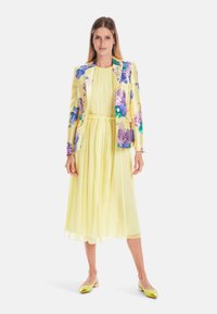Gelbes Chiffonkleid mit einem plissierten Rock, kombiniert mit einem blumenmusternden Satinblazer in Blau- und Lilatönen; gelbe Ballerinas.