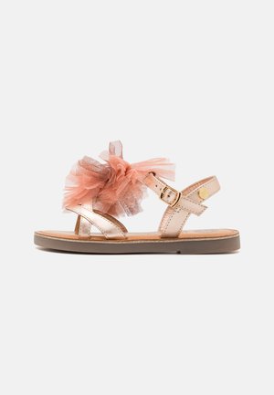 Sandaña metálica de color oro rosa con suela plana, con un gran detalle de flor de tul rosa y correa ajustable con hebilla.