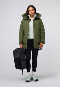 Donna che indossa un parka verde con cappuccio sopra un pile bianco, leggings neri, calze bianche e sneaker, mentre tiene uno zaino nero al lato.