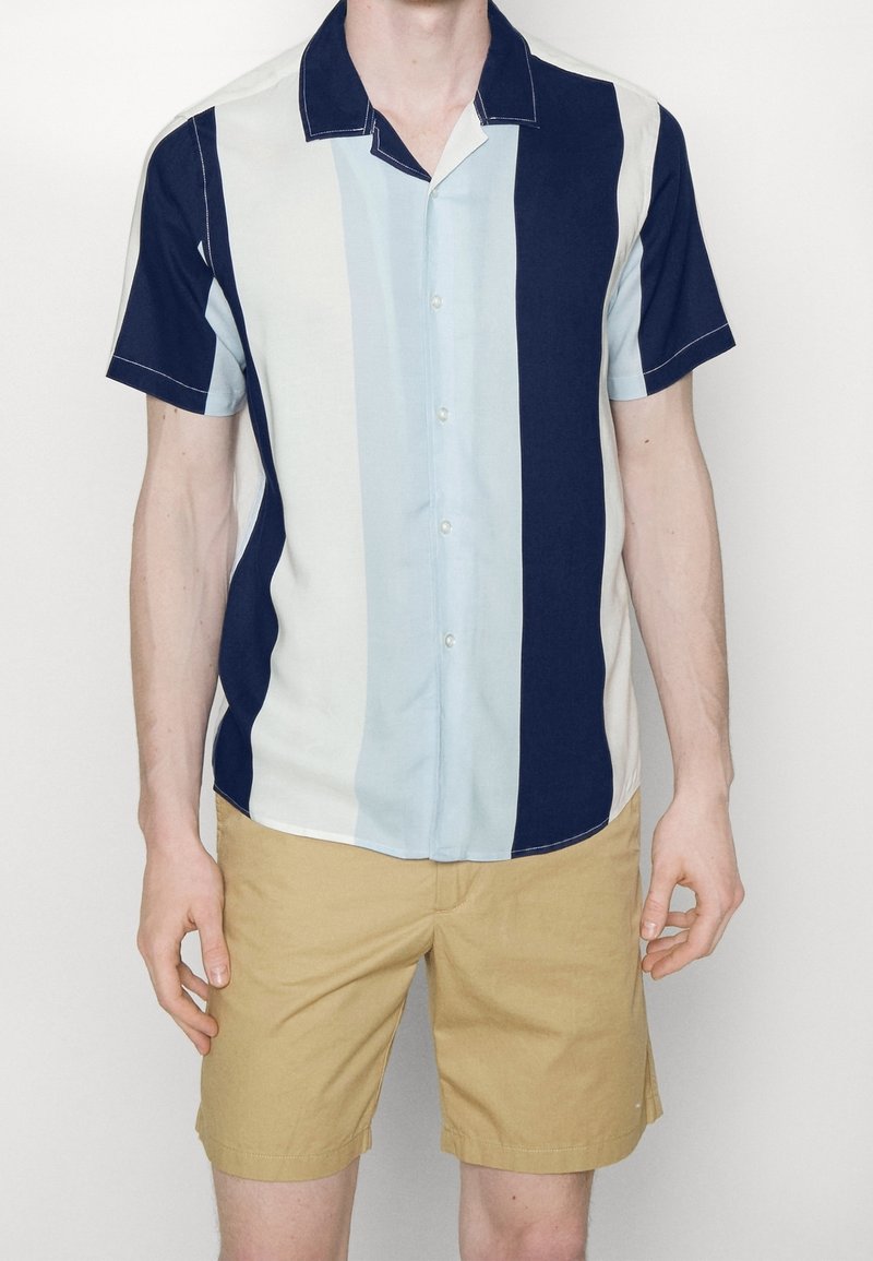 Homme portant une chemise à manches courtes à rayures verticales bleu marine, bleu clair et blanches et un short beige, debout devant un fond uni.