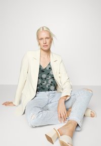 Blazer creme com lapelas de bico, blusa floral verde, jeans azul claro desfiados e sandálias de bloco em beje; um look texturizado e em camadas.