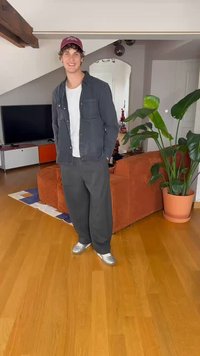 Pelēka pogu krekls virs baltas T-kreklu, pelēki bikses un sudrabaini kedas. Tumši sarkana cepure atpūšas uz cirtainiem matiem. Iekštelpu vide ar augiem.