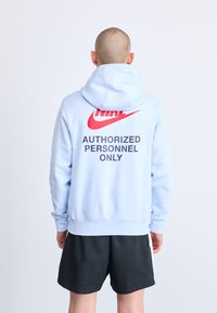 Šviesiai mėlyna džemperis su gobtuvu, su dideliu raudonu Nike logotipu ir juodu tekstu "Tik įgalioti asmenys" ant nugaros.