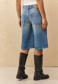 Shorts en denim Bermuda de couleur bleu clair avec un ourlet brut, comportant deux poches arrière et un patch en cuir avec logo. Associés à des bottes noires jusqu'aux genoux.
