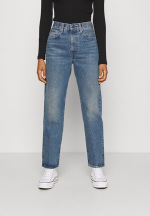 Persoon draagt een blauwe hoge taille jeans, witte sneakers en een zwarte top met lange mouwen, staand tegen een effen lichte achtergrond.