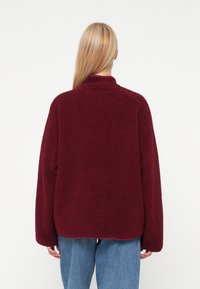 Pikkade kollaste juustega inimene kannab buršinavärvi fliisjakki ja siniseid teksaseid, seistes valge tausta poole seljaga.