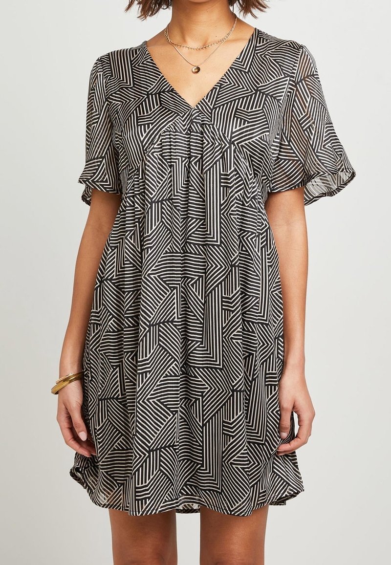 Femme portant une robe courte à motif géométrique noir et blanc, avec des manches volantées et des bracelets dorés en couches au poignet gauche.
