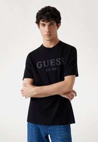 Musta puuvillapaita, jossa on harmaalla printattu "GUESS EST.1981". Lyhyet hihat ja pyöreä kaula-aukko. Yhdistetty sinisiin farkkuihin.