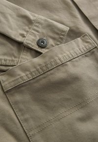 Beige cotton shirt featuring a button closure, contrast stitching, and a prominent pocket detail on the front. Smooth texture overall.
