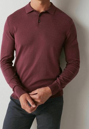 Polo shirt bordeaux à manches longues avec col, patte de boutonnage à trois boutons, poignets côtelés et texture douce. Porté avec un pantalon sombre.