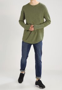 Maglione a maglia verde oliva con scollatura rotonda, abbinato a jeans in denim blu. Come calzature, indossa sneakers nere con dettagli bianchi.
