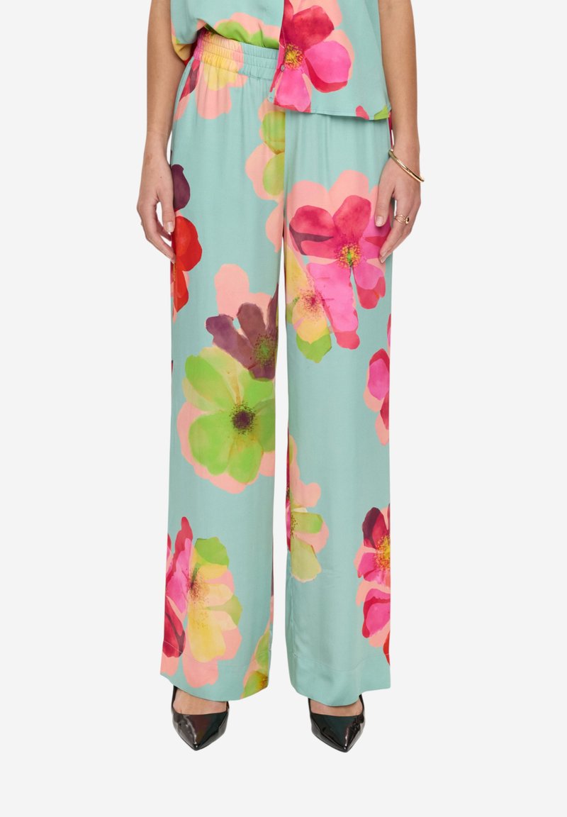Pantalon turquoise à jambes larges avec de grands motifs floraux roses, verts et rouges, porté avec des chaussures noires à bouts pointus et un haut assorti.