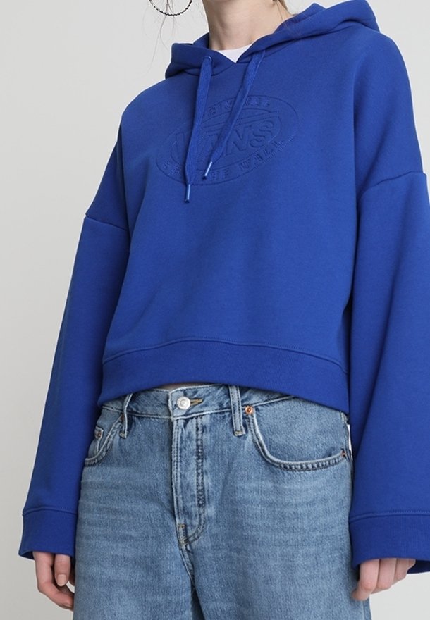 Personne portant un sweat à capuche bleu avec un logo embossé et un jean taille haute bleu clair, raccourci à la taille.