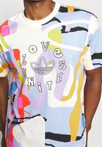 Färgglad bomullströja med abstrakta mönster i rosa, blått, lila och gult. Svart text "LOVE UNITE" med en Adidas-logga i mitten.