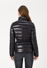 Svart pufferjakke med blank overflate, quiltet design, høy krage og et stoffbelte i midjen. Brukt sammen med grå jeans.