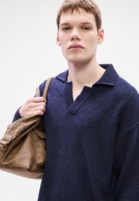 Θαλασσί πλεκτό πουλόβερ με ριμπ κολάρο και χαμηλωμένους ώμους, συνδυασμένο με καφέ δερμάτινη τσάντα που έχει φερμουάρ και πλαϊνή τσέπη.