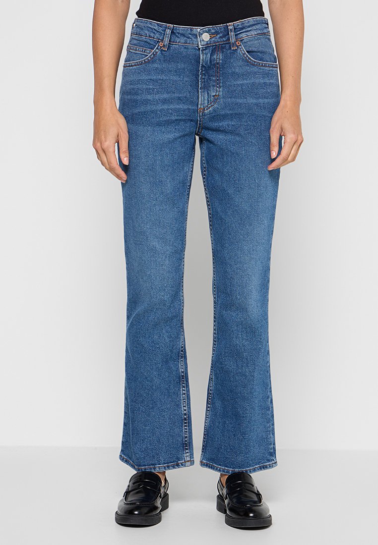 Rechtdoorgaande blauwe spijkerbroek gedragen met zwarte loafers en een zwart topje, van de taille tot de voeten weergegeven tegen een effen achtergrond.