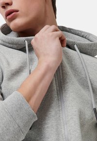 Grijze ritssluiting hoodie met een zachte textuur, voorzien van een trekkoordkap en elastische boorden. De rits is centraal aan de voorkant, wat het ontwerp versterkt.