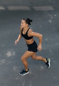 Femeie alergând pe o suprafață pavate, îmbrăcată cu un sutien sport negru, pantaloni scurți negri și adidași negri cu tălpi albe.