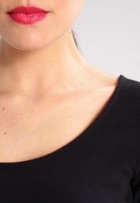 Schwarzes, geripptes Oberteil mit weit ausgeschnittenem Hals, das eine glatte Hautfarbe und kräftige, pinke Lippen zeigt und minimale Details des Schlüsselbeinbereichs zur Geltung bringt.