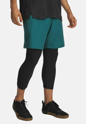 Uomo che indossa pantaloncini verdi acqua sopra leggings neri corti e scarpe da ginnastica nere, in piedi contro uno sfondo semplice.