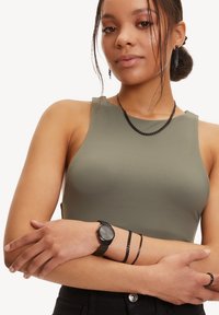 Top senza maniche verde oliva, orologio nero con cinturino testurizzato, abbinato a bracciale e collana a catena nera, modella con capelli lunghi.