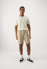 Pantalones cortos marrones con bolsillos laterales y cintura con cordón, combinados con una camiseta de manga corta color beige claro, zapatillas negras y calcetines blancos.