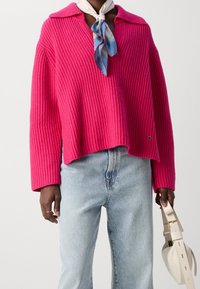 Różowy sweter o oversize'owym kroju z prążkowanej dzianiny i kołnierzykiem, zestawiony z jasnoniebieskimi dżinsami i białą torebką. Posiada luźne rękawy oraz detal z logo.