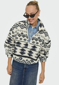 Puli od flisa u krem boji s crnim geometrijskim uzorcima, s polu-zip dizajnom i spuštenim rukavima. Uparen s traper košuljom i suknjom.